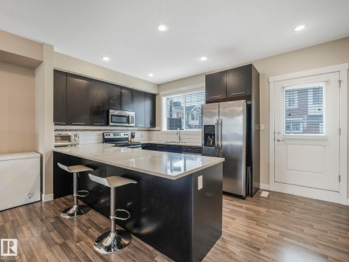 Contemporary kitchen featuring dark wood-finish cabinetry, stainless steel appliances, light-toned countertops, a full white subway tile backsplash, and wood-finish flooring - 96 8315 180 Avenue, Edmonton, AB - Indoor Photo Showing Kitchen With Stainless Steel Kitchen With Upgraded Kitchen