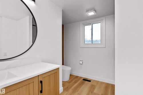 Modern bathroom featuring a wood-finish vanity with a white countertop and integrated sink, a black-framed circular mirror, and a contemporary wall-mounted light fixture - 3208 130 Avenue, Edmonton, AB - Indoor