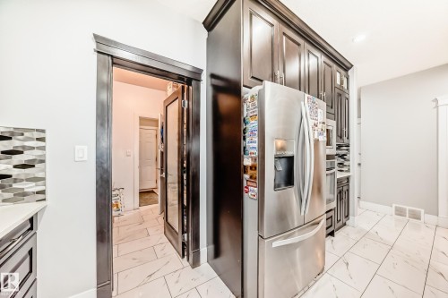 Kitchen featuring dark wood cabinetry, stainless steel appliances, a geometric tile backsplash, and light-toned tile flooring - 69 Caragana Way, Fort Saskatchewan, AB 