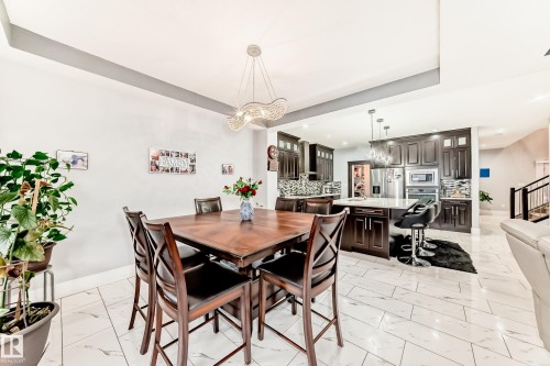 Open-concept living area featuring a formal dining space with a contemporary chandelier, a kitchen with dark wood cabinetry, a center island with pendant lighting, stainless steel appliances, and white tile flooring - 69 Caragana Way, Fort Saskatchewan, AB 