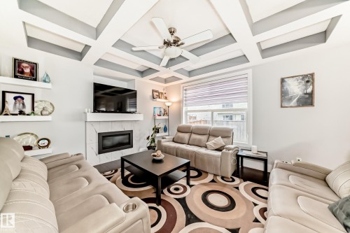 Living area featuring a coffered ceiling with a ceiling fan, a fireplace with a marble-finish surround and white mantle, dark wood-finish flooring, and a large window with horizontal blinds - 69 Caragana Way, Fort Saskatchewan, AB 