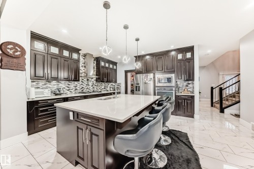 Modern kitchen featuring dark wood-finish cabinetry, a large center island with a light-toned countertop, stainless steel appliances, and a geometric tile backsplash - 69 Caragana Way, Fort Saskatchewan, AB 