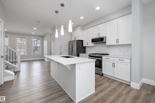 Modern kitchen featuring a white island with a sink and pendant lighting, white cabinetry, stainless steel appliances, and wood-finish flooring - 4235 Chichak Close, Edmonton, AB - Indoor Photo Showing Kitchen With Upgraded Kitchen