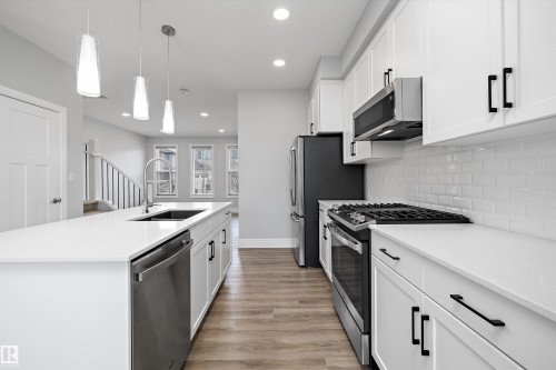 Modern kitchen featuring an expansive island with a built-in sink and stainless-steel dishwasher, white shaker cabinetry with matte black hardware, white subway tile backsplash, and wood-finish flooring - 4235 Chichak Close, Edmonton, AB - Indoor Photo Showing Kitchen With Upgraded Kitchen