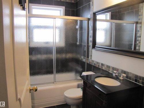 Bathroom featuring dark tile flooring and coordinating wall tiling, a white bathtub with sliding textured glass doors, and a dark wood vanity with a black countertop and oval sink - 7135 87 Street, Edmonton, AB - Indoor Photo Showing Bathroom