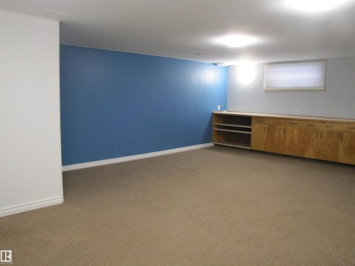 Spacious room featuring a deep blue accent wall, neutral carpeting, and a built-in wood-finish cabinet system with open shelving - 7135 87 Street, Edmonton, AB - Indoor Photo Showing Other Room