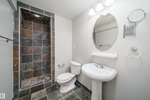 Walk-in shower featuring multi-toned slate tile, a coordinating slate tile floor, a pedestal sink with chrome faucet, an oval vanity mirror, and three-light vanity fixture - 50 14603 Miller Boulevard, Edmonton, AB - Indoor Photo Showing Bathroom