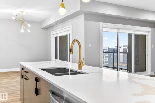 Kitchen island featuring a white countertop with integrated sink, brass-finish faucet, and wood-finish cabinetry with matte black hardware - 520 7463 May Common, Edmonton, AB - Indoor Photo Showing Kitchen With Double Sink