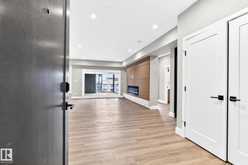 Spacious living area featuring wood-finish flooring, a contemporary fireplace with wood paneling surround, recessed lighting, and sliding glass doors opening to an outdoor space - 520 7463 May Common, Edmonton, AB - Indoor Photo Showing Other Room With Fireplace