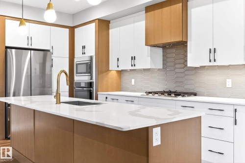 Modern kitchen featuring a large central island with an undermount sink and brass-finish faucet, white cabinetry, wood-finish accent cabinetry, stainless steel appliances, and a geometric tile backsplash - 520 7463 May Common, Edmonton, AB - Indoor Photo Showing Kitchen With Upgraded Kitchen