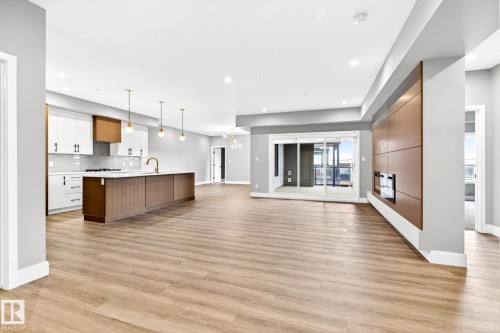 Spacious open-concept interior featuring light wood-finish flooring, a modern kitchen with a large island and pendant lighting, a built-in electric fireplace with wood paneling, and sliding glass doors leading to a balcony - 520 7463 May Common, Edmonton, AB - Indoor Photo Showing Kitchen