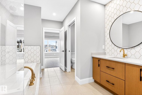 Bathroom featuring a light wood-finish vanity with a white countertop and gold-tone faucet, a round wall mirror, and hexagonal wall tiling - 520 7463 May Common, Edmonton, AB - Indoor Photo Showing Bathroom