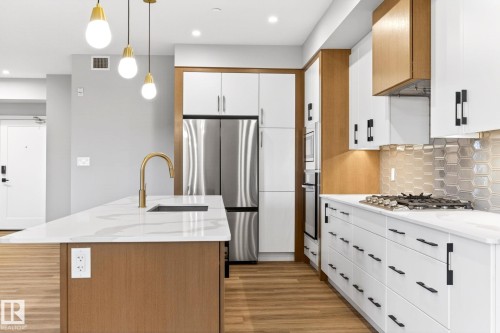 Modern kitchen featuring an island with a gold-tone faucet and undermount sink, stainless steel appliances, wood-finish flooring, white cabinetry with black hardware, and a geometric tile backsplash - 520 7463 May Common, Edmonton, AB - Indoor Photo Showing Kitchen With Stainless Steel Kitchen With Upgraded Kitchen