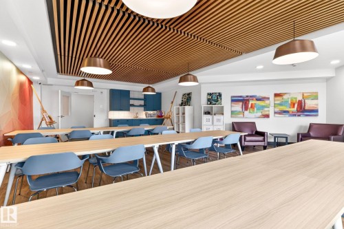 Spacious activity room featuring a slatted wood ceiling, multiple light wood-finish tables, blue chairs, and a kitchenette with blue cabinetry - 520 7463 May Common, Edmonton, AB - Indoor