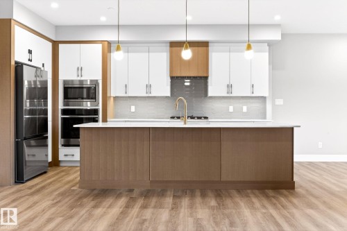 Modern kitchen featuring a large central island with a light-colored countertop, stainless steel appliances, wood-finish flooring, recessed lighting, and pendant fixtures - 520 7463 May Common, Edmonton, AB - Indoor Photo Showing Kitchen