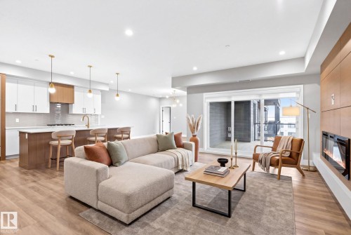 Open-concept living space featuring wood-finish flooring, a contemporary kitchen with white and wood-finish cabinetry, a large island with seating, and a built-in electric fireplace with wood paneling - 520 7463 May Common, Edmonton, AB - Indoor Photo Showing Living Room With Fireplace
