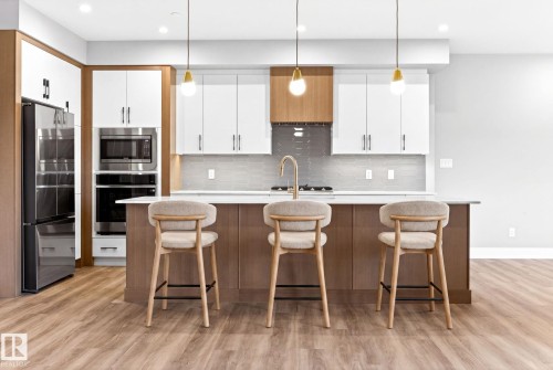 Open-concept kitchen featuring an expansive center island with seating, stainless steel appliances, and wood-finish flooring - 520 7463 May Common, Edmonton, AB - Indoor Photo Showing Other Room