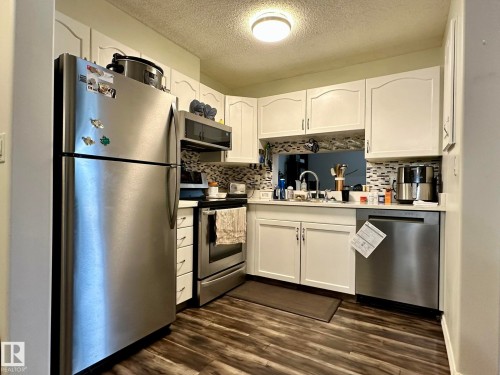 Kitchen featuring white cabinetry, stainless steel appliances, a mosaic tile backsplash, wood-finish flooring, and a pass-through window - 211 9619 174 Street, Edmonton, AB - Indoor Photo Showing Kitchen With Stainless Steel Kitchen