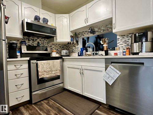 Kitchen featuring white cabinetry, stainless steel appliances, a mosaic tile backsplash, wood-finish flooring, and a pass-through window - 211 9619 174 Street, Edmonton, AB - Indoor Photo Showing Kitchen With Stainless Steel Kitchen