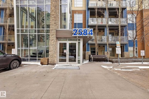 Building exterior featuring a glass-enclosed lobby, stone-clad facade elements, blue and tan siding, multi-level balconies with glass railings, and a prominent entry canopy - 409 2584 Anderson Way, Edmonton, AB - Outdoor With Balcony With Facade