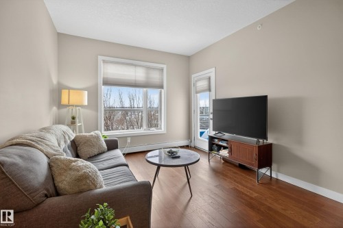 Spacious living area featuring rich wood-finish flooring, a large window providing ample natural light, neutral wall colors, and a built-in media console - 409 2584 Anderson Way, Edmonton, AB - Indoor Photo Showing Living Room