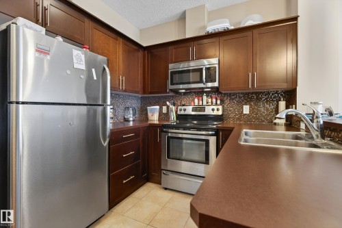 Kitchen featuring wood-finish cabinetry, stainless steel appliances, mosaic tile backsplash, and a double-basin sink - 409 2584 Anderson Way, Edmonton, AB - Indoor Photo Showing Kitchen With Stainless Steel Kitchen With Double Sink