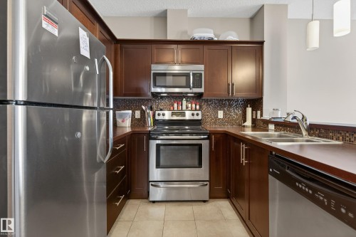 U-shaped kitchen featuring dark wood cabinetry, stainless steel appliances, a mosaic tile backsplash, and light tile flooring - 409 2584 Anderson Way, Edmonton, AB - Indoor Photo Showing Kitchen With Stainless Steel Kitchen With Double Sink