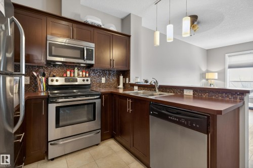 Kitchen featuring dark wood-finish cabinetry, stainless steel appliances, a mosaic tile backsplash, a double basin sink, and a breakfast bar with pendant lighting - 409 2584 Anderson Way, Edmonton, AB - Indoor Photo Showing Kitchen With Stainless Steel Kitchen With Double Sink