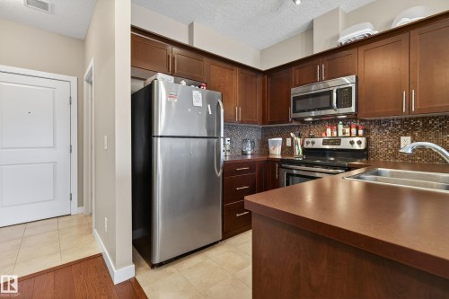 Kitchen featuring dark wood cabinetry, stainless steel appliances, a mosaic tile backsplash, and a double basin sink - 409 2584 Anderson Way, Edmonton, AB - Indoor Photo Showing Kitchen With Stainless Steel Kitchen With Double Sink