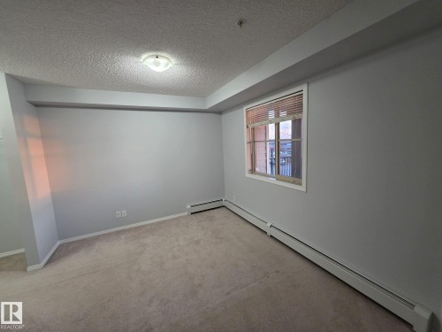 Carpeted room with light grey walls, a single window with blinds, and white baseboard heating - 310 12045 22 Avenue, Edmonton, AB 