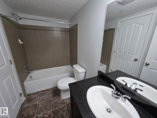 Bathroom featuring a white bathtub with tile surround, toilet, and vanity with an integrated sink and dark countertop - 310 12045 22 Avenue, Edmonton, AB 