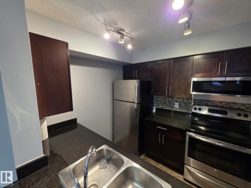 Kitchen featuring dark wood cabinetry, stainless steel appliances, a mosaic tile backsplash, and dark granite-finish countertops with an undermount double-basin sink - 310 12045 22 Avenue, Edmonton, AB 