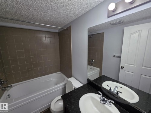 Full bathroom featuring a tub/shower combination with brown tile surround, a toilet, and a vanity with a dark countertop and undermount sink - 310 12045 22 Avenue, Edmonton, AB 