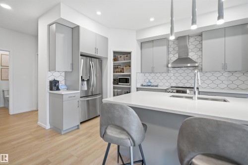 Modern kitchen featuring light wood-finish flooring, light gray cabinetry, stainless steel appliances, a geometric tile backsplash, and a large island with a white countertop and integrated sink - 7782 174A Avenue, Edmonton, AB - Indoor Photo Showing Kitchen With Upgraded Kitchen