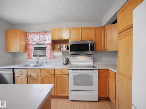 Kitchen featuring wood cabinetry, light-toned countertops, wood-finish flooring, a stainless steel microwave, and a double-basin sink - 21379 89 Avenue Nw, Edmonton, AB - Indoor Photo Showing Kitchen With Double Sink