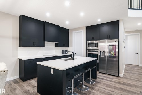 Modern kitchen featuring dark cabinetry, stainless steel appliances, a center island with bar seating, white countertops, and wood-finish flooring - 2 Edison Drive, St. Albert, AB - Indoor Photo Showing Kitchen With Double Sink