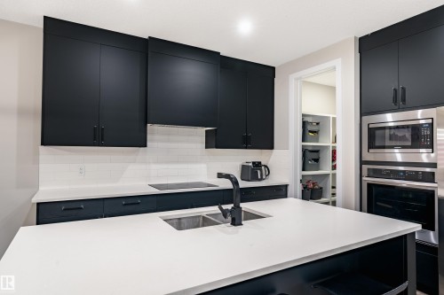 Contemporary kitchen featuring dark flat-panel cabinetry, a bright countertop island with an undermount double sink, and a stainless steel built-in microwave and oven - 2 Edison Drive, St. Albert, AB - Indoor Photo Showing Kitchen With Double Sink With Upgraded Kitchen