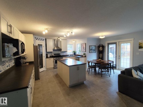 Open-concept kitchen and dining area featuring light-toned cabinetry, a central island with dark countertops, stainless steel appliances, and tile flooring - 59 Rue Monette, Beaumont, AB - Indoor