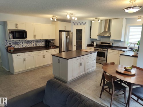 Gourmet kitchen featuring a central island with storage, white cabinetry, integrated stainless steel appliances, patterned backsplash, and tile flooring - 59 Rue Monette, Beaumont, AB - Indoor Photo Showing Kitchen With Double Sink