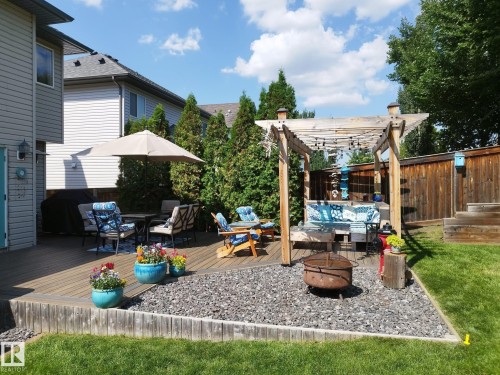 Expansive backyard featuring a multi-level wood deck, a pergola with string lighting, a dedicated gravel fire pit area, a grass lawn, and mature privacy trees - 59 Rue Monette, Beaumont, AB - Outdoor With Deck Patio Veranda With Exterior