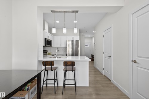 Modern kitchen featuring white shaker cabinetry, stainless steel appliances, a white subway tile backsplash, and a breakfast bar with pendant lighting - 51 603 Orchards Boulevard Sw, Edmonton, AB - Indoor Photo Showing Kitchen