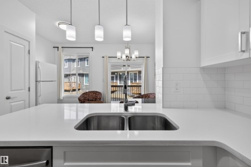 Kitchen featuring a double basin stainless steel sink, solid surface countertops, white subway tile backsplash, white cabinetry, and pendant lighting - 51 603 Orchards Boulevard Sw, Edmonton, AB - Indoor Photo Showing Kitchen With Double Sink