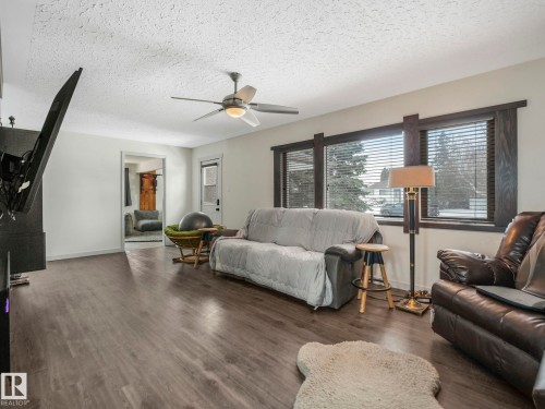 Spacious living area featuring wood-finish flooring, a ceiling fan with integrated lighting, and multiple windows with dark wood trim - 7607 70 Avenue, Edmonton, AB 