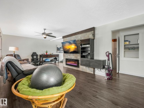 Living area featuring wood-finish flooring, a ceiling fan with integrated lighting, and a stone-look accent wall with an electric fireplace - 7607 70 Avenue, Edmonton, AB 