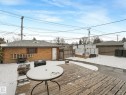 Expansive wood-finish deck with a patio table, accompanied by a brick garage with a shingle roof and a separate white door - 7607 70 Avenue, Edmonton, AB 
