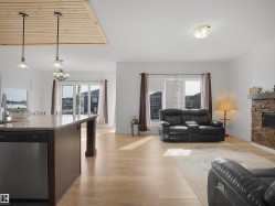 This bright interior features light-toned hardwood flooring throughout, a kitchen island with a granite countertop and integrated dishwasher, and a living area with a stone-clad fireplace - 
