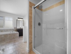 The bathroom features a tiled shower with a glass enclosure, a tiled soaking tub, and a vanity with a mirror and light fixture - 