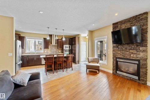 Open-concept living space featuring wood-finish flooring, a stone-clad fireplace with a built-in television mount, a kitchen island with seating, dark wood cabinetry, and stainless steel appliances - 10630 82 Street, Edmonton, AB - Indoor Photo Showing Living Room With Fireplace