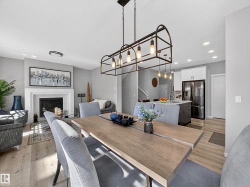Open-concept living space featuring wood-finish flooring, a contemporary linear chandelier, a fireplace with a white mantel, an island with a sink, and recessed lighting - 323 Balsam Link, Leduc, AB - Indoor Photo Showing Dining Room With Fireplace