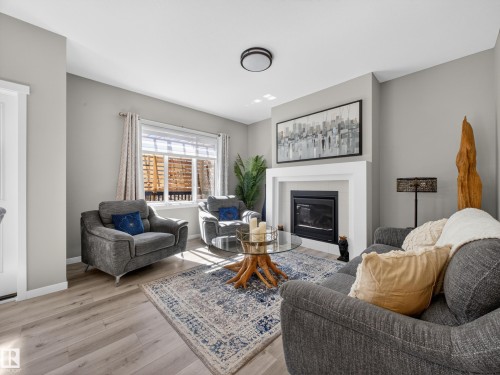 Living room featuring a fireplace with a white mantel, light gray walls, wood-finish flooring, and a window with blinds - 323 Balsam Link, Leduc, AB - Indoor Photo Showing Living Room With Fireplace